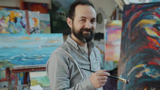 Портрет красивого бородатого художника, стоящего в студии рядом с абстрактной картиной — стоковое видео