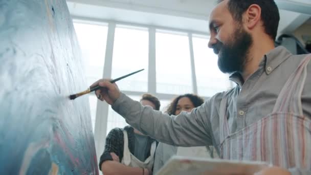 Кваліфікований дорослий художник малює картини і розмовляє зі змішаною групою студентів в студії — стокове відео