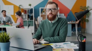 Erkek ofis çalışanının kameraya bakıp dizüstü bilgisayarla daktilo kullandığı yavaş çekim portresi.