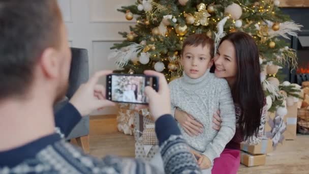 Ojciec robi zdjęcie matce i synowi w pobliżu choinki za pomocą smartfona — Wideo stockowe