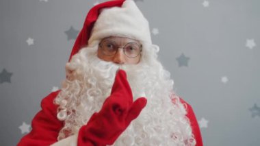 Noel Baba 'nın portresi şşşş el hareketi yapıyor ve gri yıldızlı arka plandaki kameraya bakıyor.