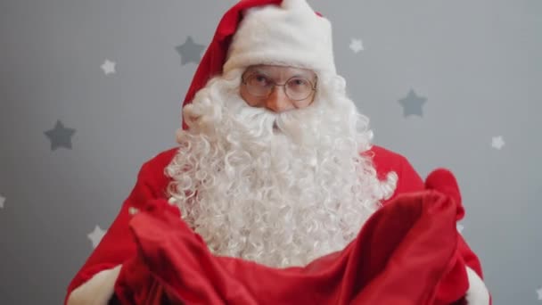 Langsom bevegelse med smilende fyr i julenissekostyme gir pose med gaver til kamera – stockvideo