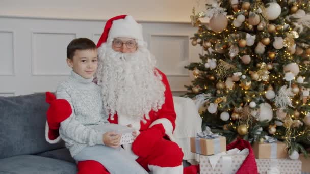 Porträt eines entzückenden Kindes mit Geschenk und Weihnachtsmann, der zu Hause auf dem Sofa sitzt und in die Kamera schaut — Stockvideo