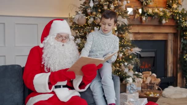 Noel Baba kostümlü bir adamın yeni yılda sevimli bir çocuğa kitap okuması. — Stok video