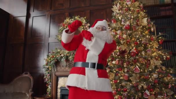 Zabawny człowiek w kostiumie Świętego Mikołaja tańczy w domu w pobliżu choinki — Wideo stockowe