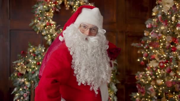 Sorrindo cara em Papai Noel terno segurando saco com presentes de pé dentro de casa — Vídeo de Stock