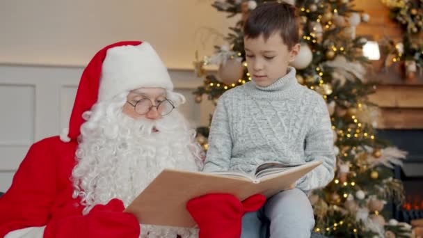 Adorable child and Santa Claus reading book near Christmas tree and talking on New Years day — Stock Video