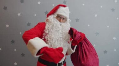 Noel Baba kıyafetli adam elinde hediyelerle yıldızlı arka plandaki kameraya bakıyor.