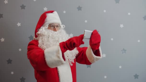 Man in Santa costume holding smartphone touching screen taking selfie on grey background — Stock Video
