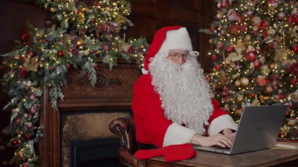 Медленное движение Санта-Клауса с ноутбуком за столом в украшенной квартире — стоковое видео