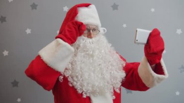 Noel Baba 'nın portresi parlak gri arka planda akıllı telefon kamerasıyla selfie çekiyor.