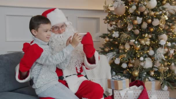 Kleiner Junge macht Selfie mit Weihnachtsmann mit neuem Smartphone in dekoriertem Zimmer — Stockvideo