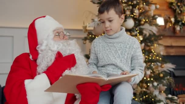 Netter Junge und Weihnachtsmann reden und lesen Buch am Neujahrstag in der Nähe geschmückter Baum — Stockvideo