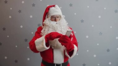 Noel Baba kostümlü ciddi bir adam gri arka planda akıllı telefon ekranı kullanıyor.