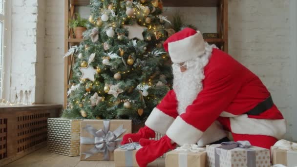 Санта Клаус у традиційному одязі ставить подарунки під ялинку у відпустку вдома. — стокове відео