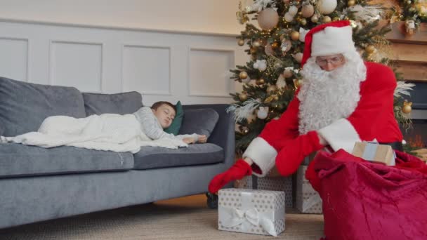 Noel Baba yeni yıl ağacının altına hediyeler yerleştirirken küçük çocuk evde uyuyor. — Stok video