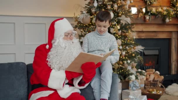 Der Weihnachtsmann liest dem glücklichen kleinen Jungen ein Buch vor und gratuliert dem Kind zu Weihnachten — Stockvideo