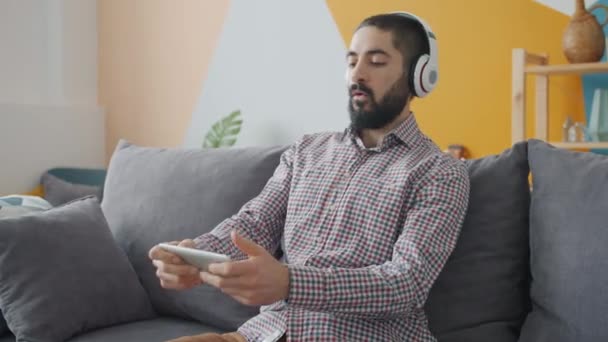 Movimento lento do homem do Oriente Médio jogando com smartphone usando fones de ouvido — Vídeo de Stock
