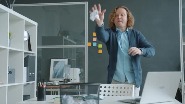 Радісний чоловік грає в гру в офісі кидаючи паперові кульки в бін розважається один — стокове відео