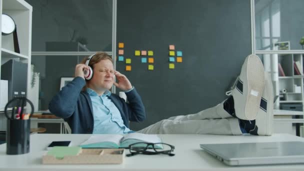 Hombre alegre disfrutando de la música en heaphones bailando brazos móviles en el escritorio en la oficina — Vídeos de Stock