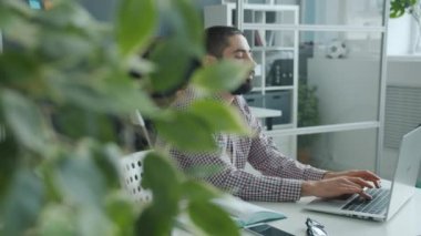 Yorgun bir ofis çalışanı dizüstü bilgisayarla yazı yazıp yüzlere dokunuyor. Baş ağrısı ve yorgunluk hissediyor.