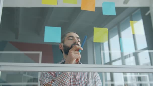 Movimiento lento del empresario de raza mixta que trabaja con notas adhesivas en la pared de vidrio en la oficina — Vídeos de Stock