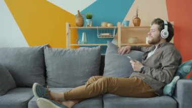 Kulaklıklı neşeli adam müzik dinliyor ve akıllı telefon kullanıyor. Evde dinleniyor.