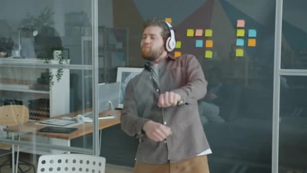 Movimiento lento de chico alegre bailando en el lugar de trabajo con auriculares inalámbricos — Vídeos de Stock