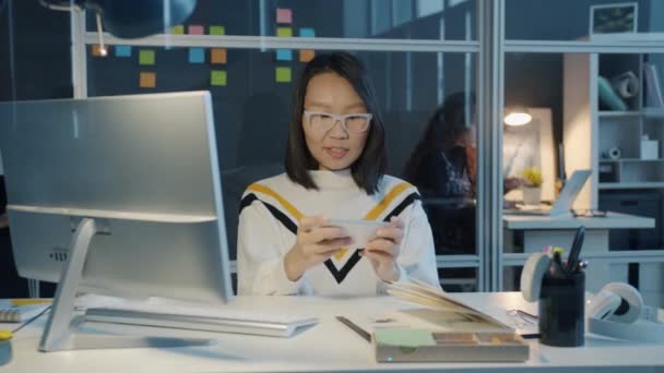 Menina asiática feliz relaxando no escritório jogando celular vídeo game tocando tela do smartphone à noite — Vídeo de Stock