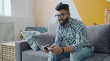 Mutlu Orta Doğulu adam evde oturup video oyunu oynuyor ve kontrolü elinde tutuyor.