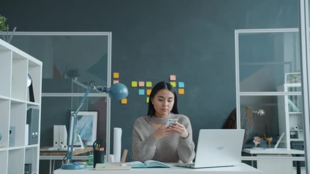 Felice ragazza asiatica utilizzando smartphone sorridente sms al lavoro mentre la donna sta lavorando in background — Video Stock