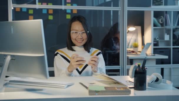 Veselé asijské obchodní dáma baví s smartphone videohry v tmavé kanceláři — Stock video