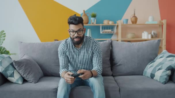 Portret van een vreugdevolle Arabische man die vido speelt met joystick en thuis naar de camera kijkt — Stockvideo