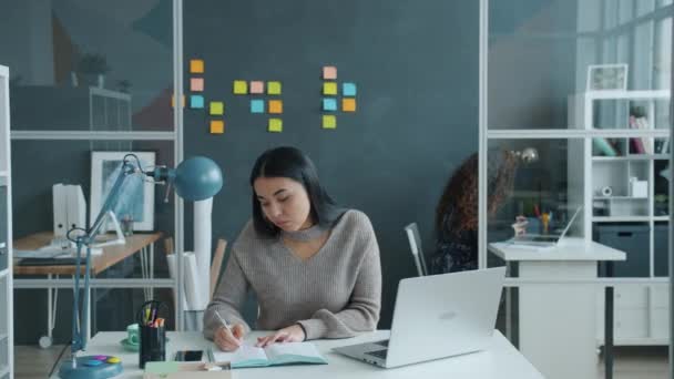 Druk aziatisch kantoor werknemer met behulp van laptop schrijven in notebook, vrouw werkt op de achtergrond — Stockvideo