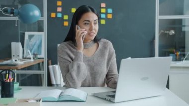 Cep telefonuyla konuşan mutlu melez kadın ofiste dizüstü bilgisayar kullanarak iyi haberler alıyor.
