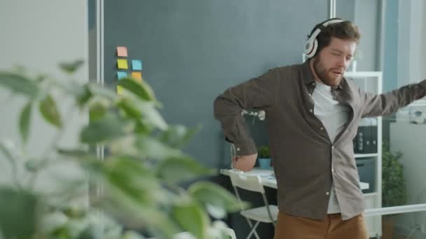 Glad ung affärsman dansar på kontoret lyssnar på musik via hörlurar — Stockvideo