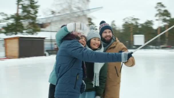 Happy men and women holding smartphone with selfie stick and taking photos in ice-skating rink — Stock Video