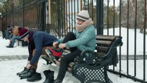 Pessoas colocando patins no gelo sentado no banco no ringue de patinação no parque de inverno — Vídeo de Stock