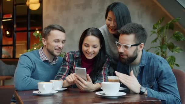 Amigos felices hablando y riendo usando un teléfono inteligente mirando la pantalla en la mesa en la cafetería — Vídeos de Stock