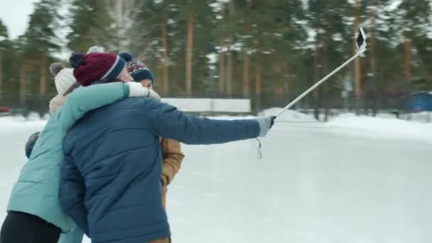 Homens e mulheres alegres tirando fotos usando selfie stick e smartphone na pista de patinação no gelo — Vídeo de Stock