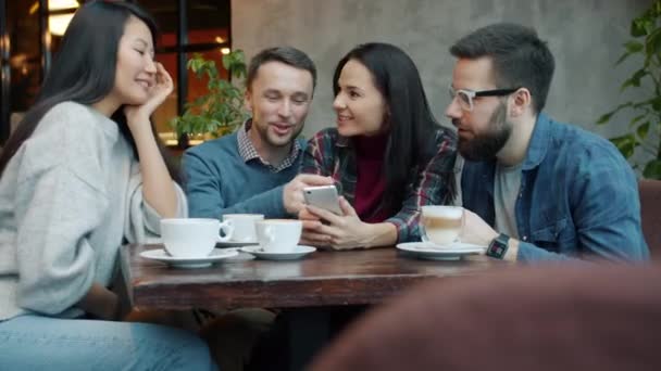 Мильтиэтническая группа друзей с помощью смартфона разговаривает и смеется в уютном кафе — стоковое видео