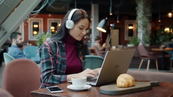 Женщина-фрилансер, работающая с ноутбуком, печатает и наслаждается музыкой с наушниками в кафе — стоковое видео