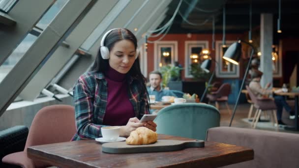 Девушка в наушниках наслаждается музыкой и использует смартфон за столом в кафе — стоковое видео