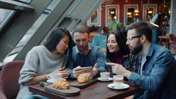 Młodzi szczęśliwi ludzie oglądają zdjęcia na smartfonie, rozmawiają i śmieją się w kawiarni — Wideo stockowe