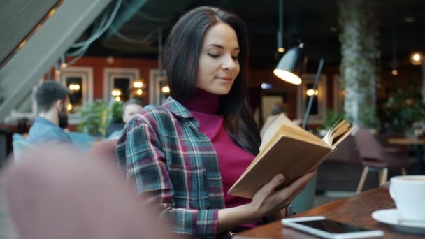 Привлекательная девушка читает интересную книгу в кафе расслабляясь сидя за столом в одиночестве переворачивая страницу — стоковое видео