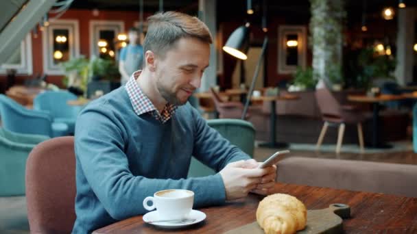 Glada ung man med hjälp av smartphone sms och leende sitter vid bordet i café — Stockvideo