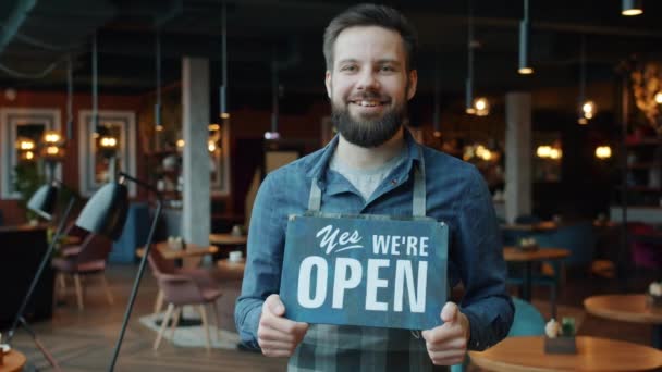 Portrait of cheerful handsome young waiter in apron holding we are open sign in coffee shop — Stock Video