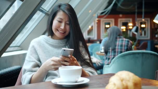 Радостная азиатка с помощью смартфона трогает экран улыбаясь и смеясь за столом в кафе — стоковое видео