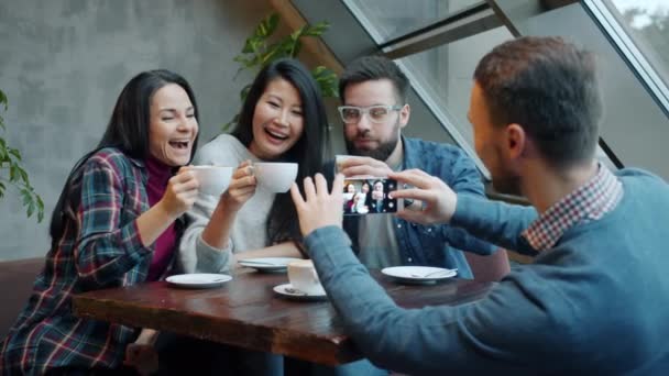 Młody człowiek robi zdjęcia szczęśliwym przyjaciołom pozującym z filiżankami kawy w nowoczesnej kawiarni — Wideo stockowe