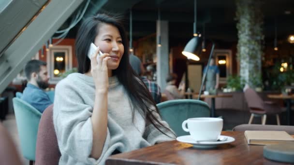 Привлекательная азиатская девушка болтает по мобильному телефону смеясь наслаждаясь разговором в кафе — стоковое видео